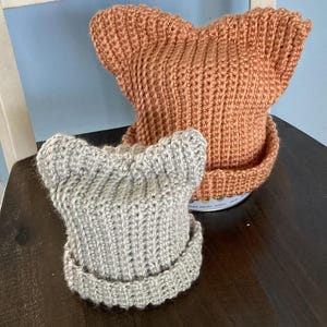 Könnte beinhalten: Zwei handgefertigte Strickmützen mit spitz zulaufenden Ohrendetails sitzen auf einem Holzstuhl. Eine Mütze ist hellgrau, die andere hat eine warme orangebraune Farbe. Die Mützen haben einen gerippten Rand und ein strukturiertes Strickmuster.