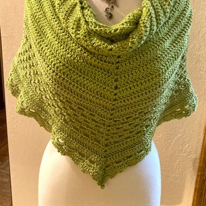 May include: A green crocheted shawl with a scalloped edge. The shawl is draped over a white mannequin with a decorative butterfly necklace.