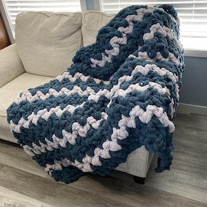 May include: A chunky knit blanket in a chevron pattern, draped over a beige sofa. The blanket features alternating rows of navy blue and light gray yarn, creating a cozy and textured appearance. The blanket is a soft home decor item.