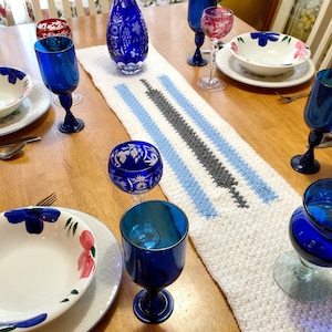 Könnte beinhalten: Ein weißer gehäkelter Tischläufer mit blauen und grauen Streifen. Der Läufer liegt auf einem Holztisch mit blauen und klaren Glaskelchen, weißen Tellern mit Blumenmustern und einer blauen Glaskaraffe.