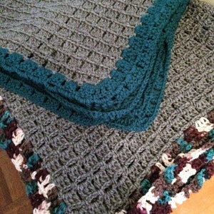 Peut inclure: Une couverture en crochet gris avec une bordure turquoise et une bordure en crochet multicolore. La couverture est réalisée avec un motif de point simple et a une texture douce et confortable.