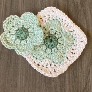 May include: Two handmade crochet items. One is a flower shape, the other is a square with a flower design. Both are in shades of white, light green, and dark green. The items are placed on a wooden surface.
