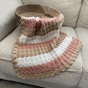 May include: A crocheted baby blanket with a scalloped edge in shades of pink, white, and beige. The blanket is draped over a wicker basket on a beige couch.