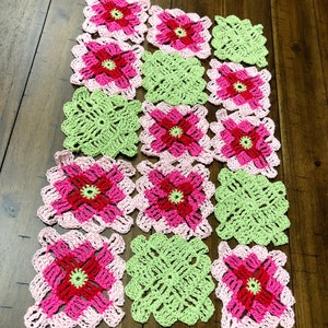 May include: A crocheted blanket made of pink, green, and red squares. The squares are arranged in a pattern and have a floral design.