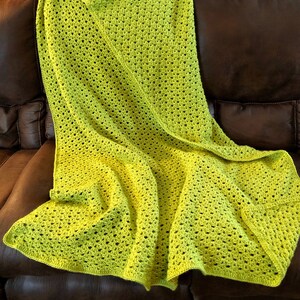 May include: A vibrant lime green crochet blanket, with an open weave design, draped over a brown leather sofa. The blanket's bright colour contrasts with the dark sofa. The blanket appears soft and inviting, perfect for a cosy evening.