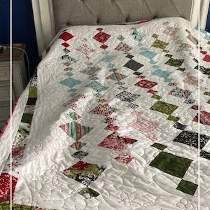 May include: A white quilt with a diamond pattern in red, green, and blue fabrics. The quilt is on a bed with a gray headboard. The quilt is quilted with a swirl pattern. The text "Patternprincess.com" is visible in the bottom right corner.