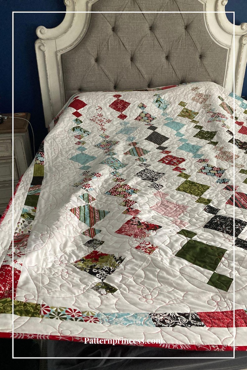 May include: A white quilt with a diamond pattern in red, green, and blue fabrics. The quilt is on a bed with a gray headboard. The quilt is quilted with a swirl pattern. The text "Patternprincess.com" is visible in the bottom right corner.