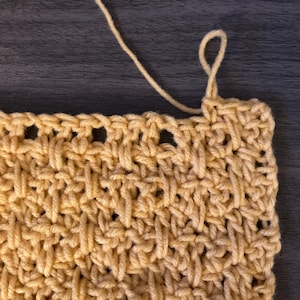 May include: A rectangular piece of light yellow yarn fabric featuring a textured basketweave crochet pattern. The fabric has a loose strand of yarn extending from the top right corner, showing the interconnected loops of the stitch work.