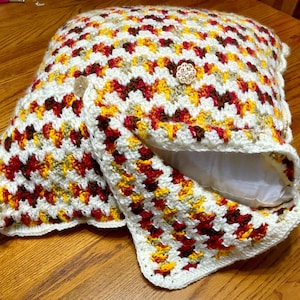 May include: A crocheted pillow cover with a geometric pattern in shades of red, yellow, brown, and white. The cover is folded over a white pillow insert.