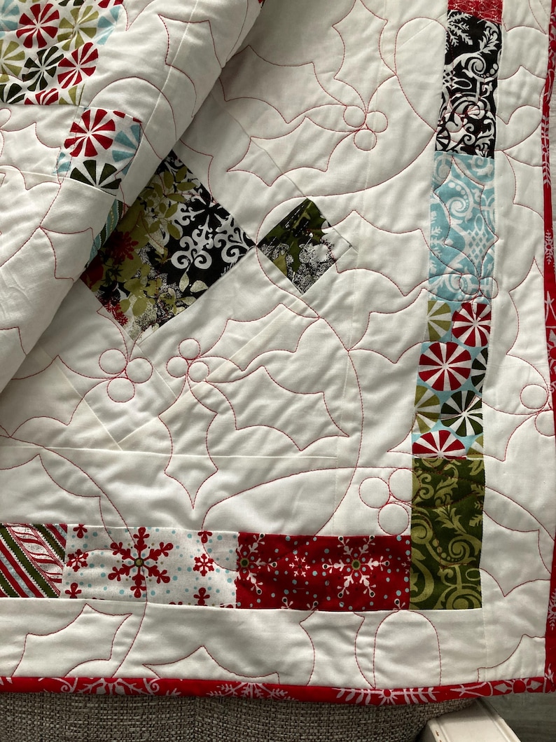 May include: A white quilt with a red and green holly leaf pattern. The quilt is made of various fabrics with different patterns, including snowflakes, candy canes, and geometric designs.