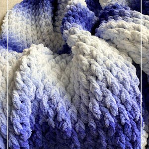 May include: A blue and white ombre chunky knit blanket with a soft, fluffy texture. The blanket is made with a thick yarn and has a loose, open weave. The blanket is folded and draped over a couch.