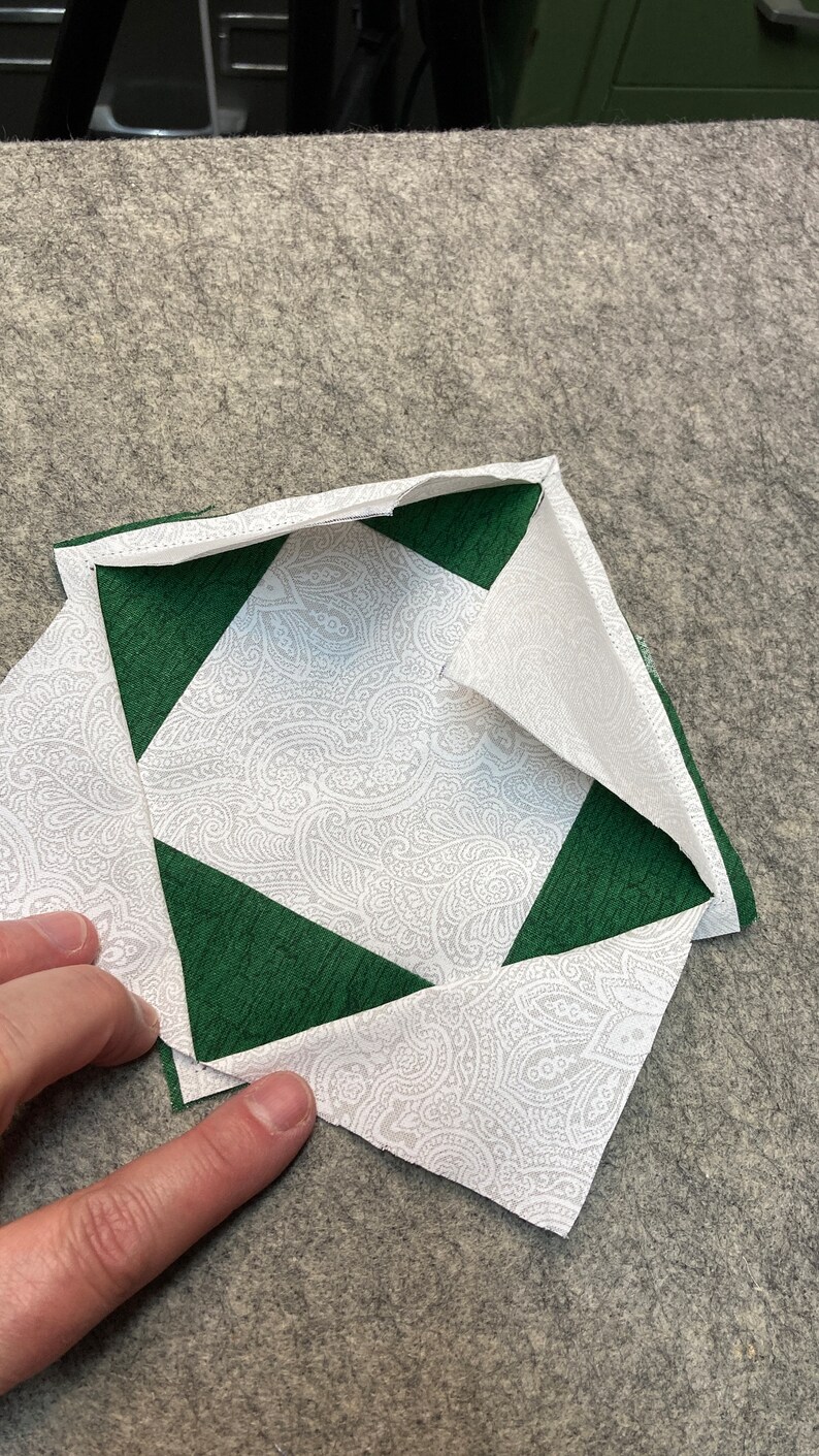 May include: A white fabric square with a paisley pattern is folded in half, with the corners of the fabric folded in to create a diamond shape. The fabric is surrounded by a green fabric border.