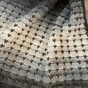 Op de afbeelding: Een handgehaakte deken in grijstinten, die overgaat van licht naar donker. De deken heeft een textuurpatroon, waardoor een visueel aantrekkelijk en gezellig ontwerp ontstaat. Het garen lijkt zacht en de deken is opgevouwen.