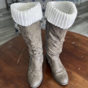 Puede incluir: Un par de botas altas marrones desgastadas con puños de bota de punto blanco. Las botas tienen un aspecto desgastado y texturizado y están sobre una superficie de madera. Los puños blancos añaden un elemento de contraste.