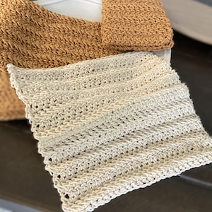 Puede incluir: Primer plano de dos paños de cocina de ganchillo. Uno es de color beige claro y el otro es marrón medio. Los paños de cocina están hechos de hilo texturizado y están doblados sobre una superficie marrón oscuro.