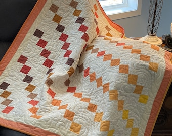 Pode incluir: Uma colcha branca com um design de patchwork com quadrados de cor laranja, castanho e vermelho. A colcha está dobrada e drapeada sobre uma poltrona cinzenta escura.