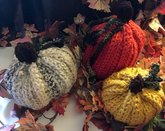 Puede incluir: Tres calabazas hechas a mano con ganchillo en crema, naranja y amarillo, cada una con un tallo marrón y acentos de vid verde. Las calabazas están dispuestas sobre un lecho de hojas de otoño, creando una exhibición de temporada.