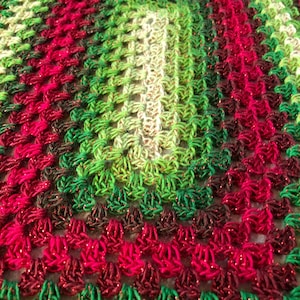 Pode incluir: Uma manta de crochê com um degradê de cores verde, vermelho e branco. A manta é feita com um fio brilhante e tem uma forma quadrada.