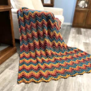 Puede incluir: Una manta de ganchillo hecha a mano con un patrón de chevrón. Presenta colores cálidos como rojo, naranja, amarillo y azul. La manta está sobre un sillón blanco, mostrando su textura y diseño.