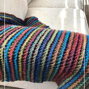 Puede incluir: Una manta de crochet a rayas multicolores drapeada sobre un sofá gris claro. La manta está hecha con una variedad de tonos de azul, verde, marrón y rojo. La manta es texturizada y tiene un patrón de puntada en forma de gofre.