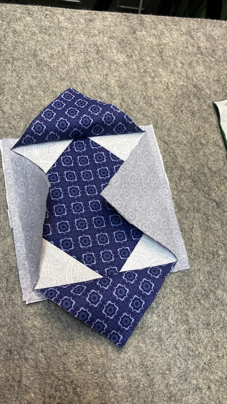 May include: A blue and white patchwork quilt block with a geometric pattern. The block is made of four triangles and a square in the center. The triangles are folded over the square, creating a diamond shape.