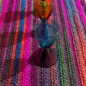 Pode incluir: Três copos com haste de vidro de diferentes cores, vermelho, âmbar e azul, estão alinhados em um corredor de mesa de crochê colorido com listras rosa, azul, verde e marrom.