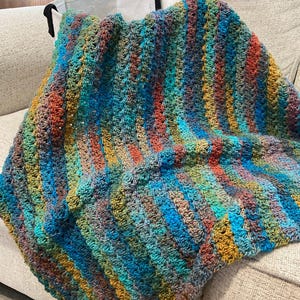May include: A handmade crochet blanket featuring a variegated yarn pattern with vertical stripes in shades of blue, green, orange, and brown. The textile has a textured, repeating stitch pattern throughout, creating a soft and thick appearance.