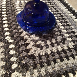 May include: A blue glass butter dish with a domed lid sits on a crocheted table runner with a black and white diamond pattern.