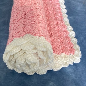 May include: A pink crocheted baby blanket with a white crocheted border. The blanket is rolled up and has a scalloped edge.