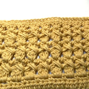 May include: A mustard yellow crocheted fabric swatch featuring a repeating textured pattern of crossed stitches and small clusters. The piece is finished with a straight, solid row of single crochet stitches along the top and bottom edges.