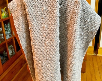 Può includere: Una morbida coperta in crochet grigia con un motivo testurizzato a punto bobble. La coperta è drappeggiata su una sedia di legno.