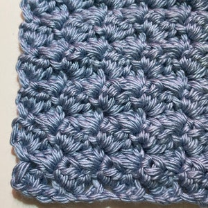 May include: A close-up of a light blue crocheted fabric with a crochet hook resting on top. The fabric has a textured, woven pattern.