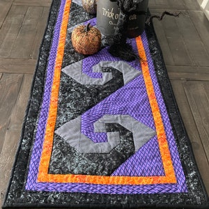 Może przedstawiać: Czarny, fioletowy i pomarańczowy bieżnik na stół na Halloween z geometrycznym wzorem. Bieżnik przedstawia czarne drzewko z drutu z napisem "Trick or Treat" na podstawie.