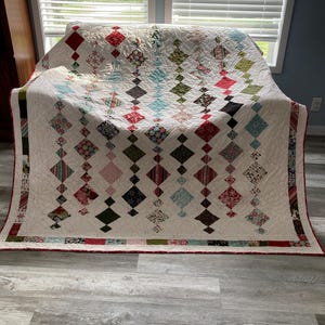 May include: A white quilt with a diamond pattern in shades of red, green, blue, and black. The quilt is folded and sitting on a light gray wood floor.
