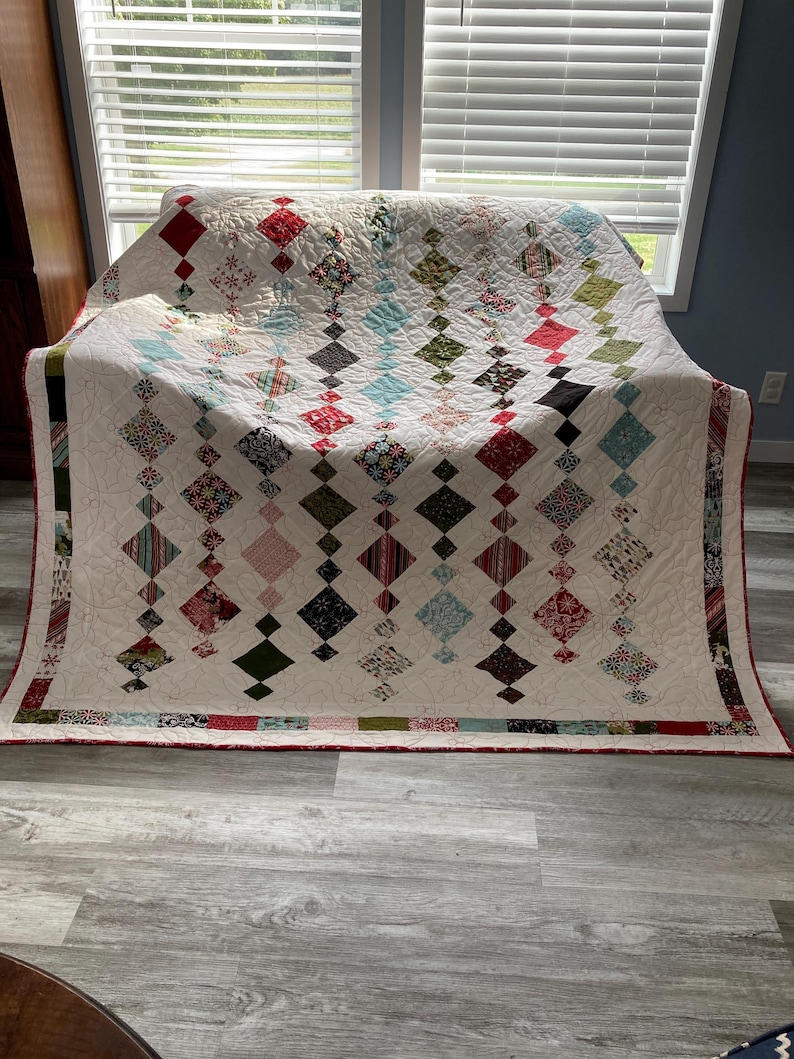 May include: A white quilt with a diamond pattern in shades of red, green, blue, and black. The quilt is folded and sitting on a light gray wood floor.