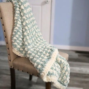 May include: A soft, textured blanket in shades of light blue and cream, draped over a beige upholstered chair. The blanket features a repeating pattern of puffy, textured squares and a cream-colored border. The blanket is a cozy home decor item.