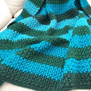 May include: A handmade, striped blanket in shades of teal and forest green. The blanket is made of a thick, textured yarn, with horizontal stripes of varying widths. The blanket is draped over a light-colored surface.