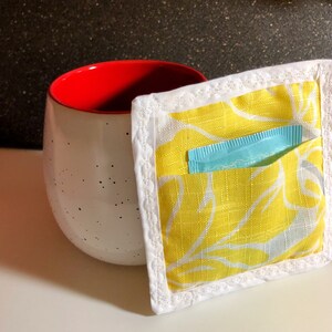 May include: A white mug with red rim and black speckles, next to a yellow fabric pouch with a white floral pattern and a blue packet inside. The pouch has a white border.
