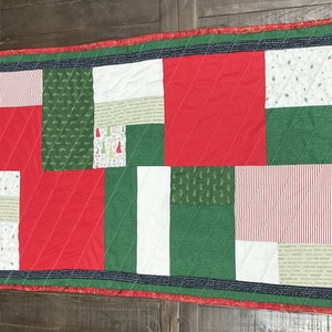 May include: A red, green, and white patchwork quilt with a Christmas theme. The quilt is made of various fabrics with different patterns, including stripes, polka dots, and snowflakes.