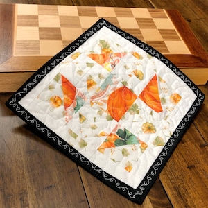 Op de afbeelding: Een witte, gestikte katoenen onderzetter met een zwarte gestikte rand. De onderzetter heeft een patchwork-ontwerp met oranje pompoenen, groene bladeren en bloemmotieven.