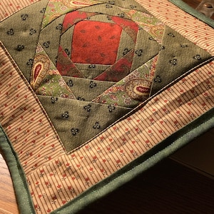 May include: A green, red and brown quilted square coaster with a paisley pattern in the centre. The coaster has a green border.