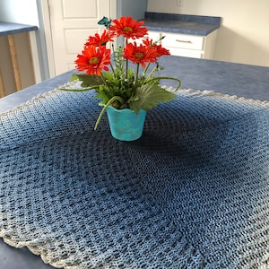 May include: A blue and white crocheted table runner with a white crocheted border. A blue ceramic pot with orange flowers sits on the table runner.