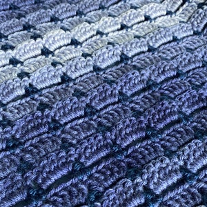May include: A close-up of a crocheted blanket with a gradient of blue and purple hues. The blanket is made with a textured stitch that creates a raised pattern.