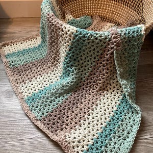 May include: A crocheted baby blanket in shades of brown, white, and teal. The blanket is made with a simple stitch pattern and has a fringed edge.