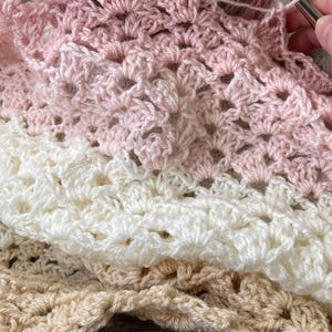 May include: A close-up of a crochet project in progress, featuring a gradient yarn in shades of pink, brown, and white. The crochet work displays a textured pattern, with a crochet hook visible, indicating the crafting process. The project is a handmade item.