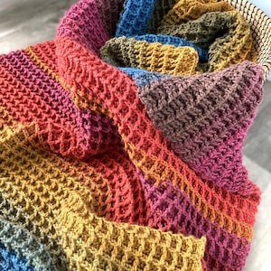 Puede incluir: Una manta colorida, tejida a mano con un patrón de punto de gofre. La manta presenta un vibrante degradado de colores, incluyendo tonos de azul, amarillo, naranja, rosa y morado. La manta está doblada y colocada en una cesta tejida.