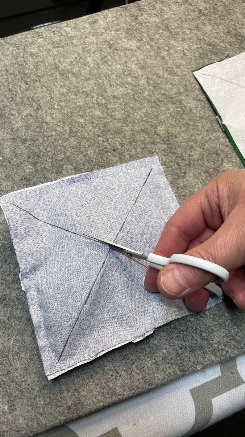 May include: A white fabric square with a gray and white pattern is marked with lines for cutting. A hand holds a pair of white scissors to cut the fabric.
