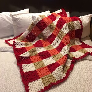 May include: A crocheted blanket with a checkered pattern in red, white, orange, and brown. The blanket is folded and draped over a white bed.