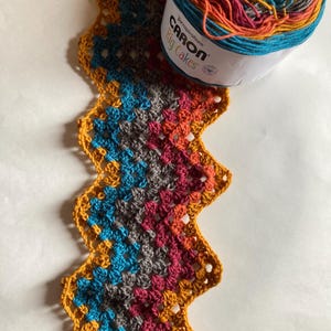 May include: A crocheted scarf in a zig-zag pattern using a multi-colored yarn with shades of gray, teal, orange, and red. The yarn is from Caron Big Cakes yarn brand.