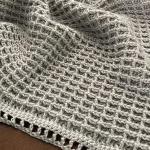 Peut inclure: Une couverture au crochet gris clair avec un motif en forme de gaufre. La couverture est pliée, montrant la texture et le design ajouré. Le bord a une bordure décorative avec de petites ouvertures.